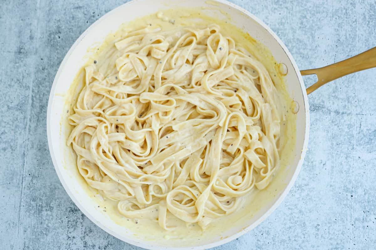 High protein chicken Alfredo fettuccine pasta coated in a creamy white sauce sits in a large white pan with a wooden handle, placed on a light gray textured surface.