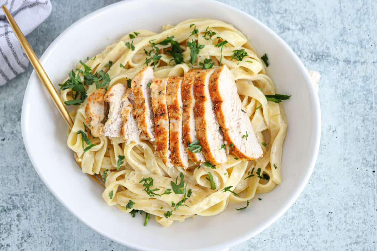 A white bowl filled with high protein chicken alfredo, featuring fettuccine topped with sliced grilled chicken breast and garnished with chopped parsley. A gold fork rests on the side of the bowl.