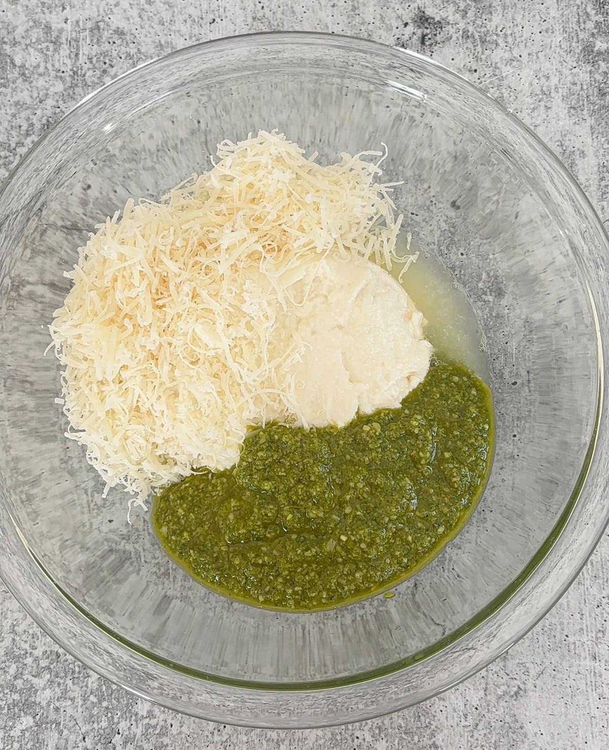 A glass bowl contains three ingredients for ricotta pesto pasta: shredded cheese, creamy white sauce, and vibrant green pesto, arranged separately on a gray countertop.