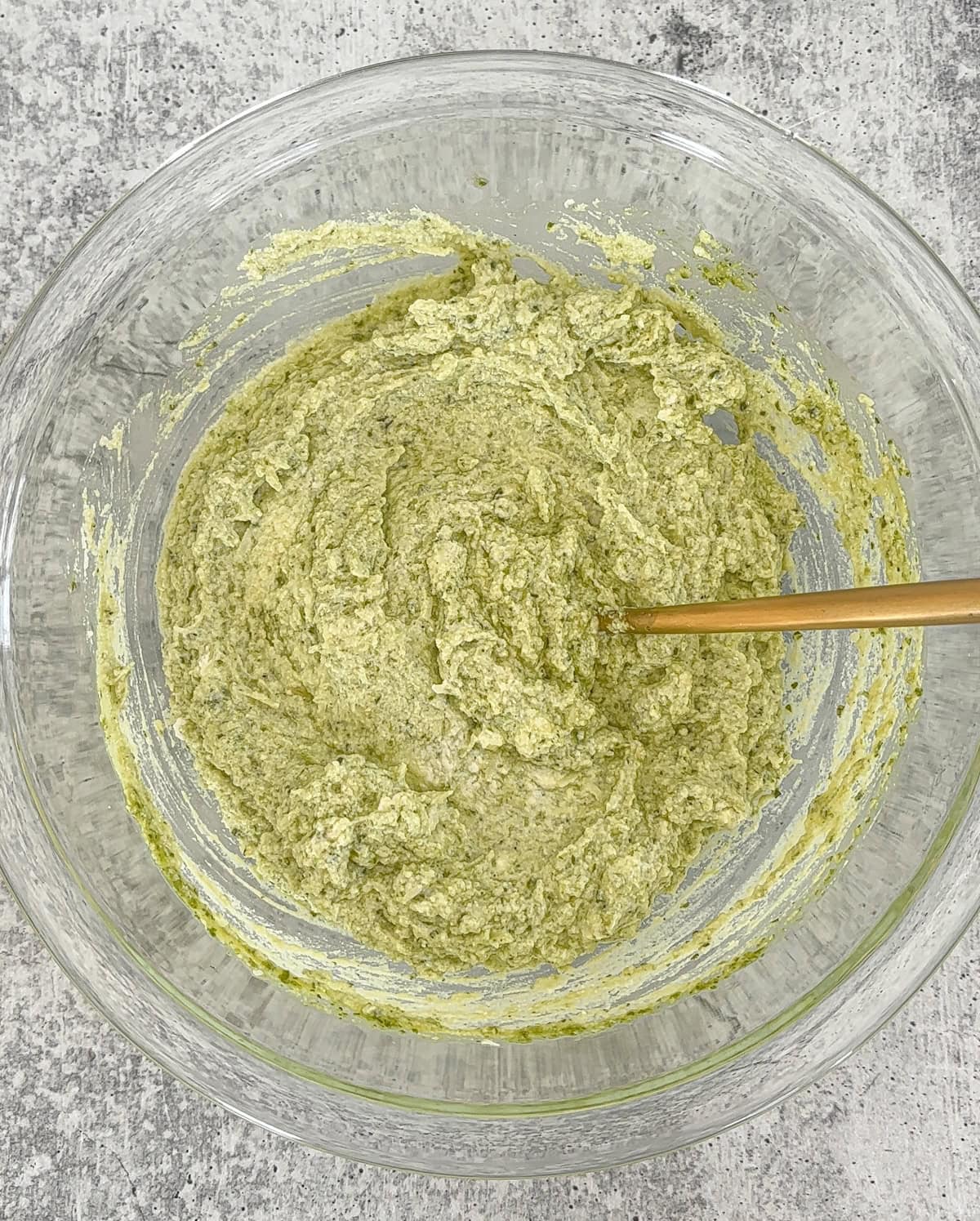 A glass bowl containing a thick, pale green ricotta pesto pasta mixture being stirred with a wooden spoon, set on a light gray textured surface.