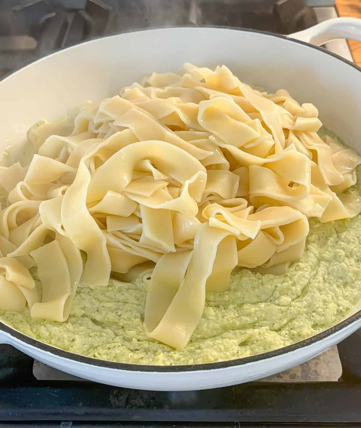 Wide, flat pappardelle pasta sits atop a creamy ricotta pesto sauce in a white pan on a stovetop, ready to be stirred together. Steam rises from the hot dish.