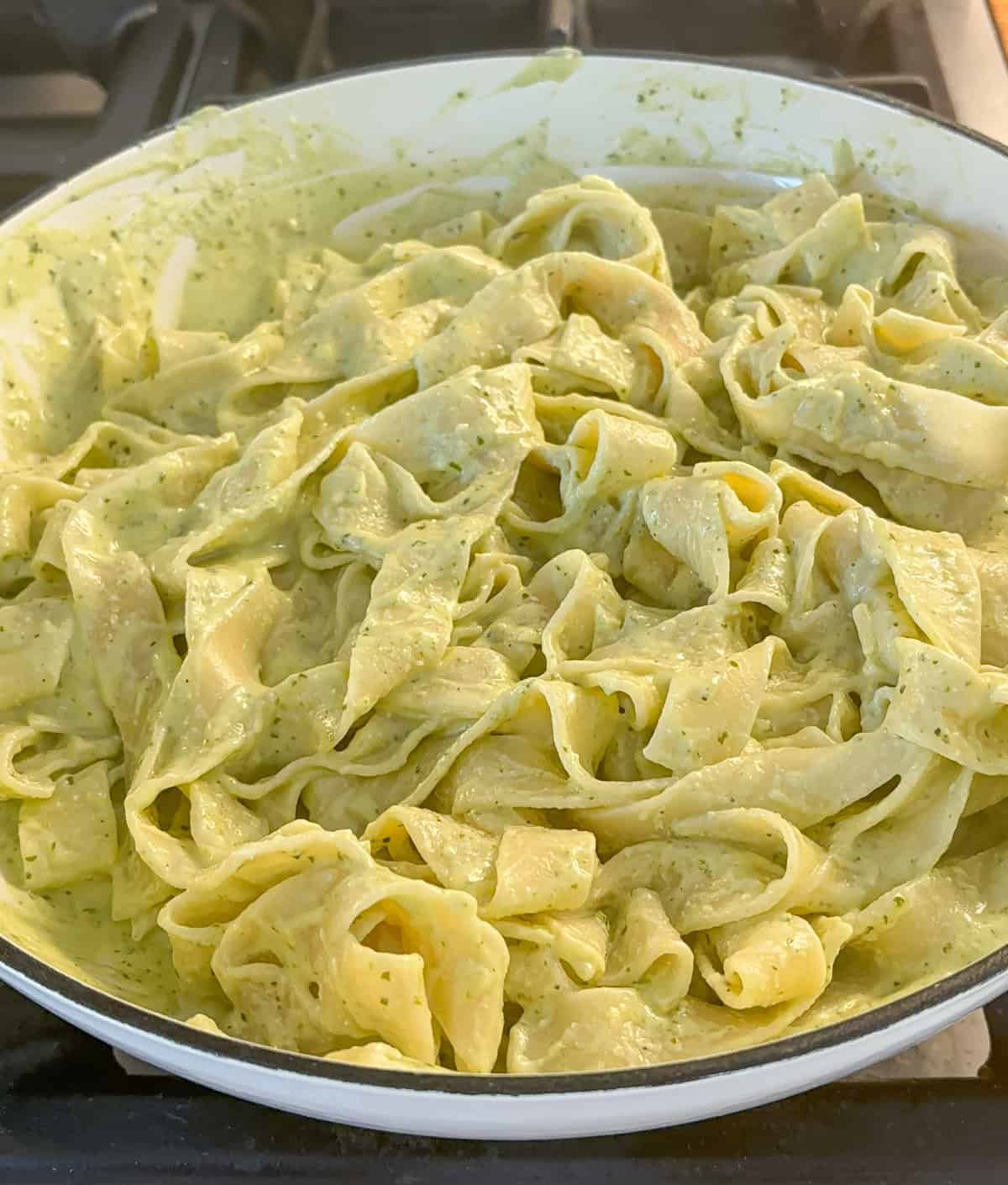 A pan filled with wide, ribbon-like ricotta pesto pasta coated in a creamy green sauce, sitting on a stovetop.