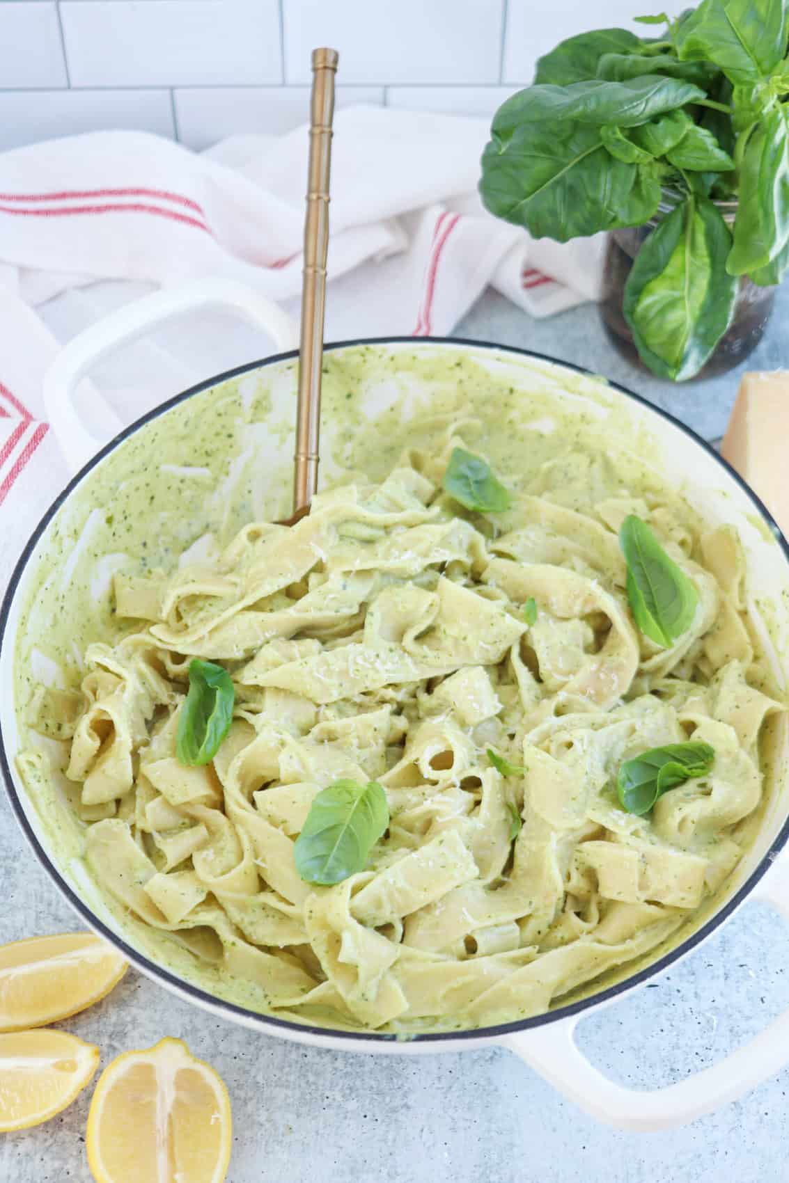 A white pot filled with creamy ricotta pesto pasta, coated in green pesto sauce and garnished with fresh basil leaves. Lemon wedges and a bunch of basil are nearby on a light countertop. A gold fork rests in the pasta.