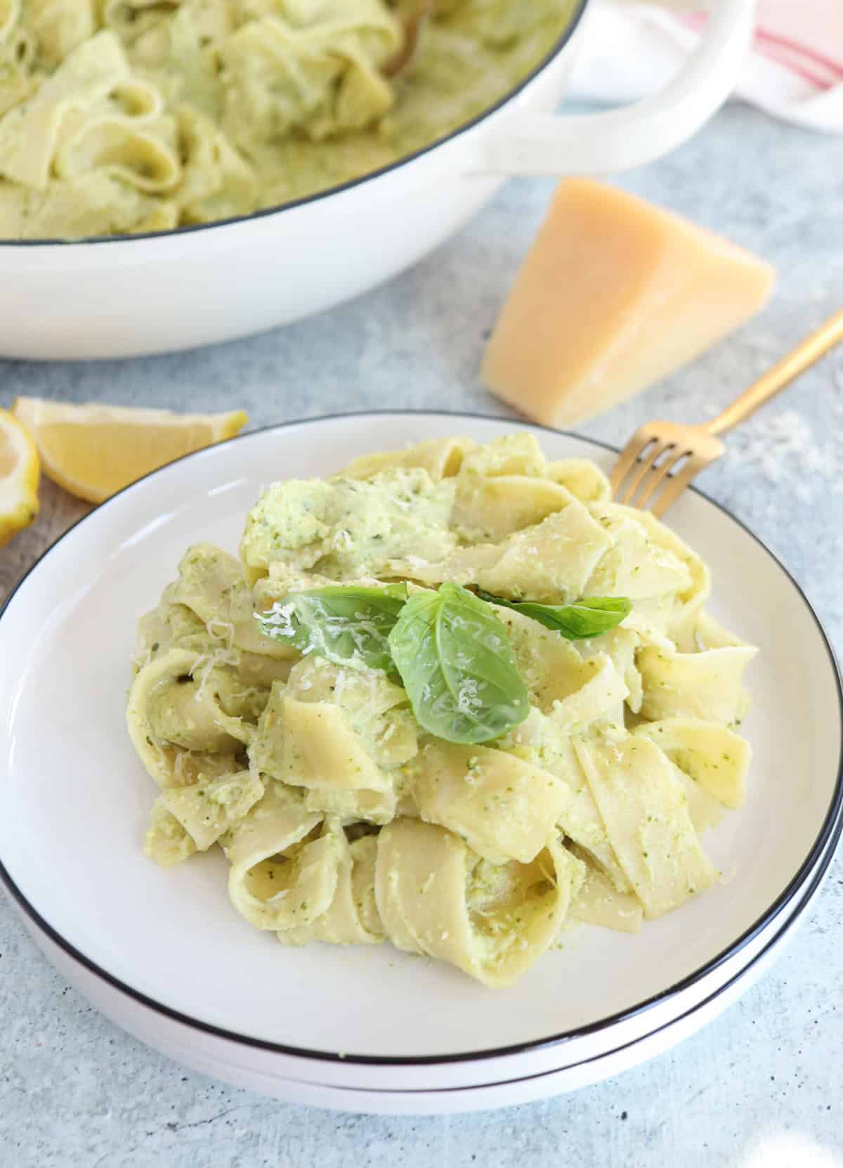 A plate of wide pappardelle coated in creamy ricotta pesto pasta sauce, garnished with fresh basil leaves. A wedge of cheese, a lemon slice, and a fork are nearby, with more pasta in a white pan in the background.