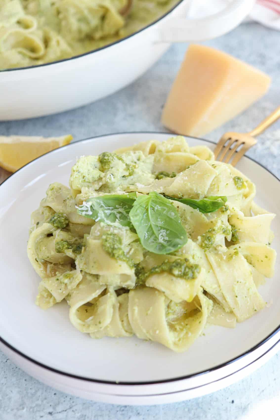 A plate of wide ricotta pesto pasta, coated in green pesto sauce and garnished with fresh basil and grated cheese, sits beside a wedge of Parmesan cheese and a gold fork on a light surface.