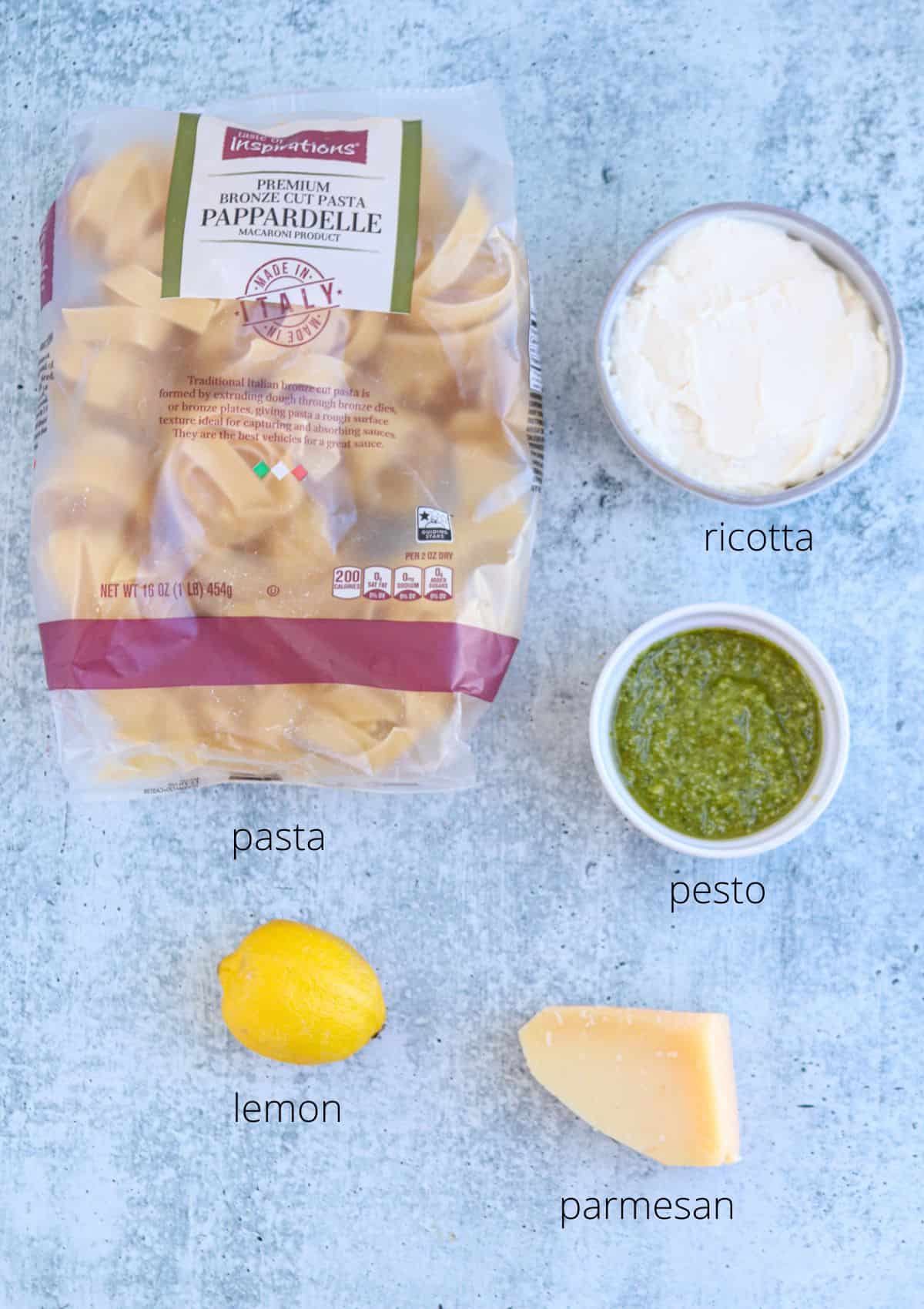 A package of pappardelle pasta, a bowl of creamy ricotta pesto, a lemon, and a wedge of parmesan are arranged on a light blue surface, each labeled with its name.