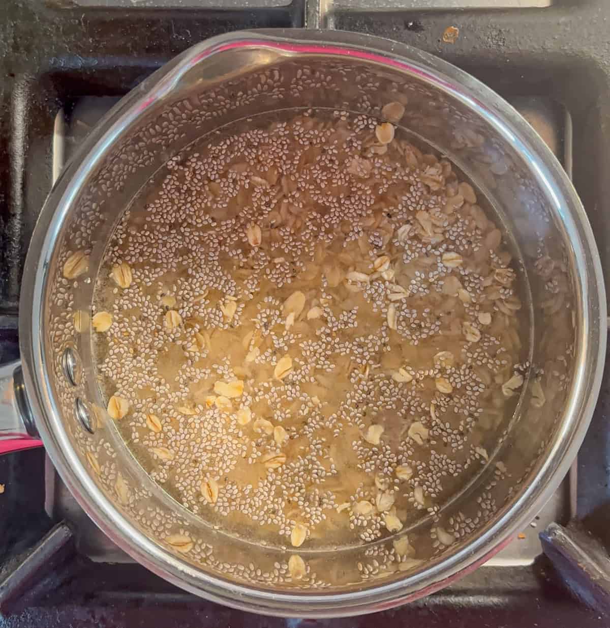 A pot on a stovetop contains water mixed with oats and chia seeds, seen from above. The grains and seeds are partially floating, much like pasta in boiling water for a ricotta pesto pasta dish, with burners visible around the pot.