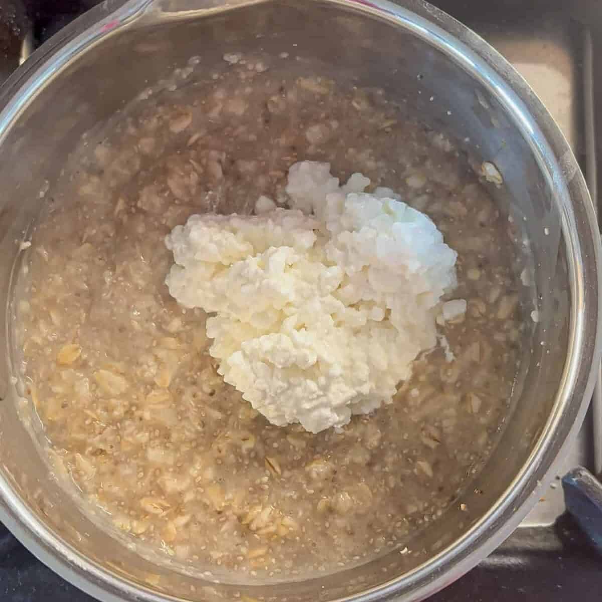 A pot of oatmeal cooking on a stove, with a dollop of cottage cheese on top, yet to be stirred in, brings comfort much like a bowl of warm ricotta pesto pasta.