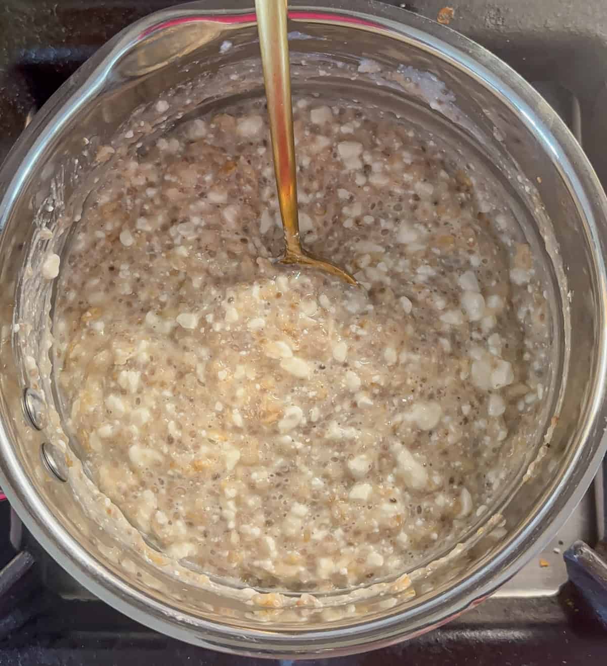 A pot of oatmeal mixed with cottage cheese is shown on a stovetop, resembling the creamy, textured look of ricotta pesto pasta. A spoon stirs the thick, lumpy mixture with visible grains and curds.