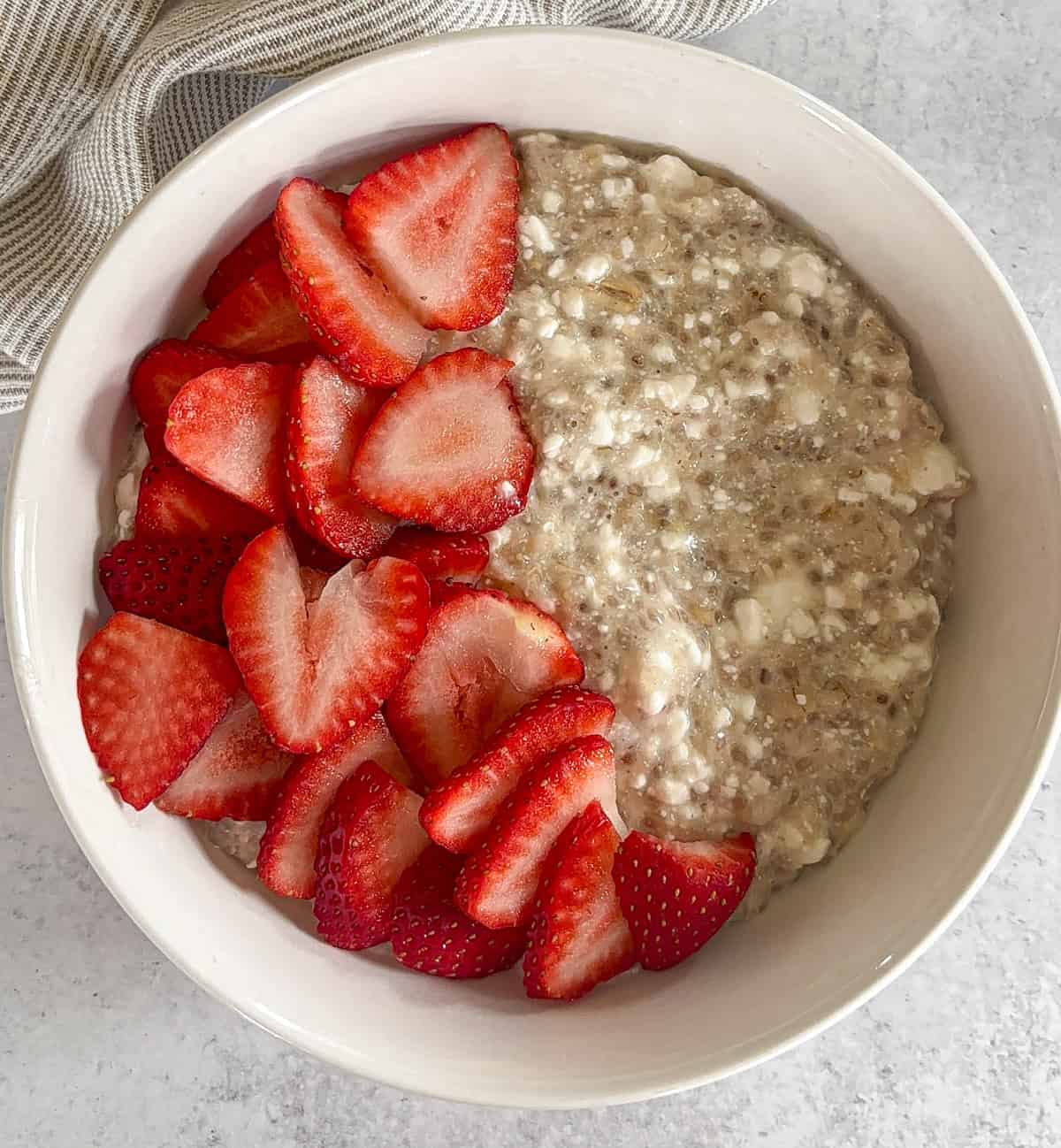 A bowl of oatmeal topped with fresh, sliced strawberries on one side sits on a light surface with a striped cloth in the background—perfect beside your favorite ricotta pesto pasta for a balanced meal.