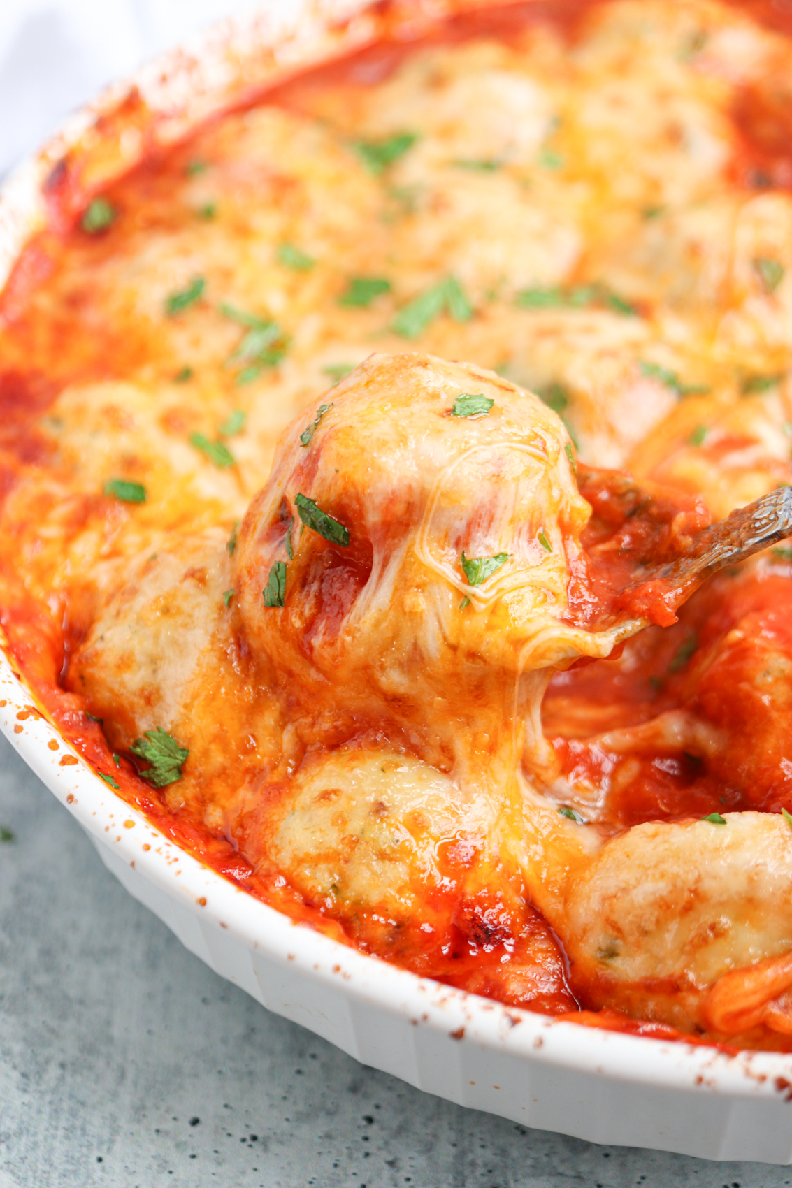 Baked cheesy chicken parm meatballs in tomato sauce, topped with melted cheese and garnished with chopped parsley, in a white dish. A spoon is lifting a cheesy meatball from the dish.