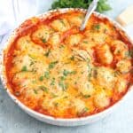 A round baking dish filled with cheesy, golden-brown baked chicken meatballs in tomato sauce, topped with melted cheese and sprinkled with chopped parsley.