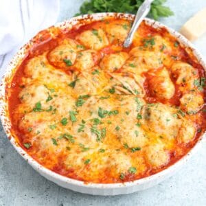 A round baking dish filled with cheesy, golden-brown baked chicken meatballs in tomato sauce, topped with melted cheese and sprinkled with chopped parsley.