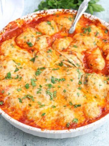 A round baking dish filled with cheesy, golden-brown baked chicken meatballs in tomato sauce, topped with melted cheese and sprinkled with chopped parsley.