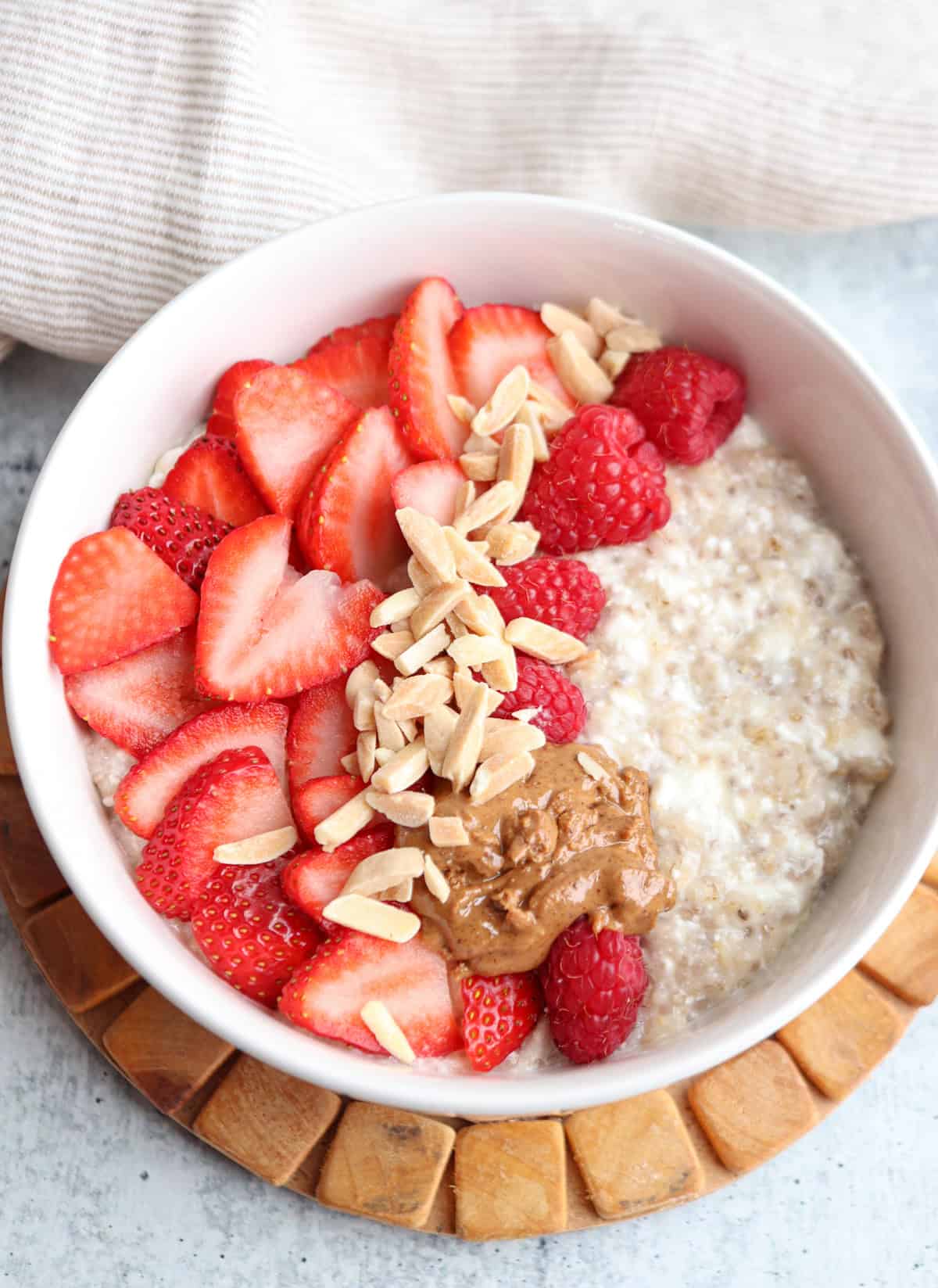A bowl of cottage cheese oatmeal topped with fresh strawberry slices, raspberries, slivered almonds, and a dollop of almond butter, placed on a wooden coaster—perfect before savoring a hearty ricotta pesto pasta later in the day.