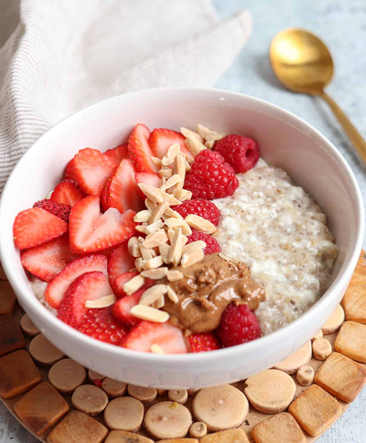 A bowl of oatmeal topped with sliced strawberries, raspberries, almond butter, and slivered almonds sits on a wooden mat, alongside a gold spoon and beige napkin—an inviting scene reminiscent of relaxed ricotta pesto pasta mornings.
