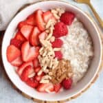 A bowl of cottage cheese oatmeal topped with sliced strawberries, raspberries, slivered almonds, and a dollop of nut butter sits next to a gold spoon and striped napkin—a cozy breakfast just as comforting as ricotta pesto pasta.