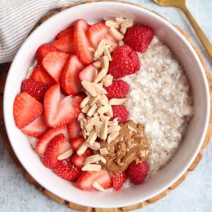 A bowl of cottage cheese oatmeal topped with sliced strawberries, raspberries, slivered almonds, and a dollop of nut butter sits next to a gold spoon and striped napkin—a cozy breakfast just as comforting as ricotta pesto pasta.