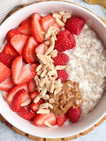 A bowl of cottage cheese oatmeal topped with sliced strawberries, raspberries, slivered almonds, and a dollop of nut butter sits next to a gold spoon and striped napkin—a cozy breakfast just as comforting as ricotta pesto pasta.