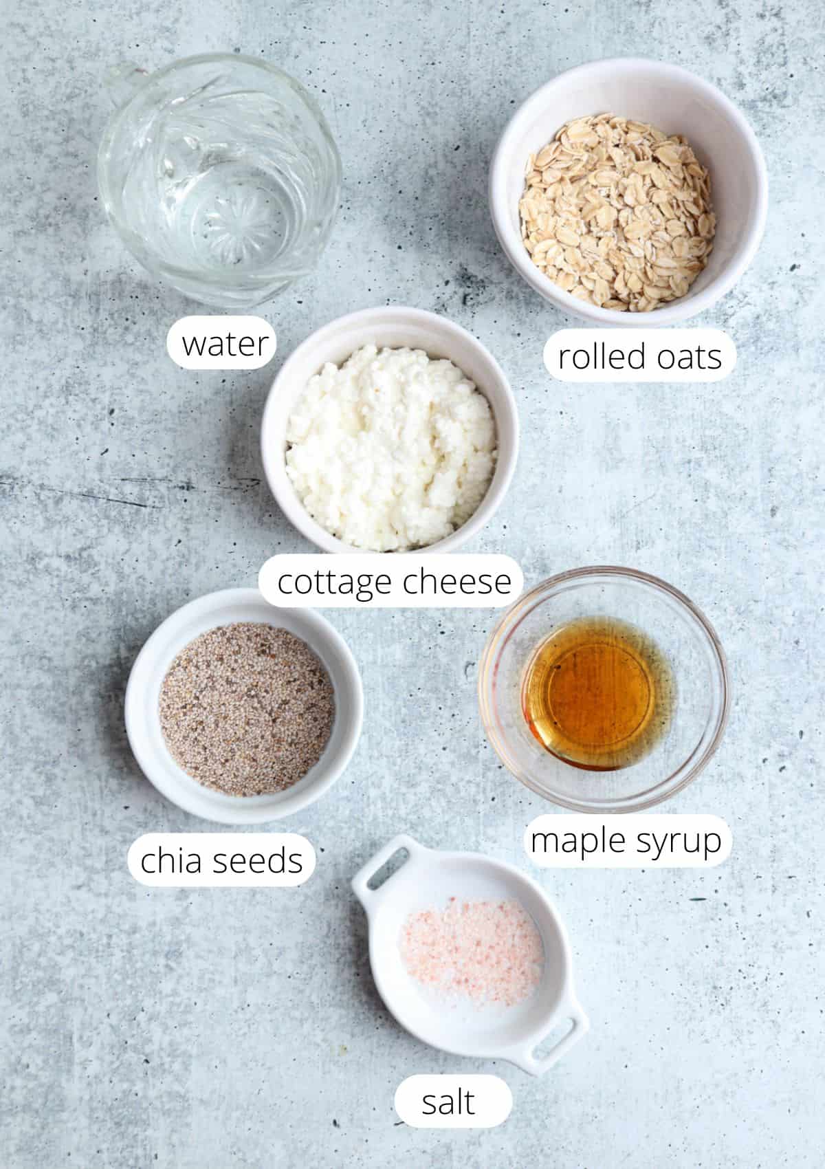 Six labeled bowls on a gray surface containing water, rolled oats, cottage cheese, chia seeds, maple syrup, and salt—similar to prepping for ricotta pesto pasta, these ingredients are arranged separately with clear labels next to each.