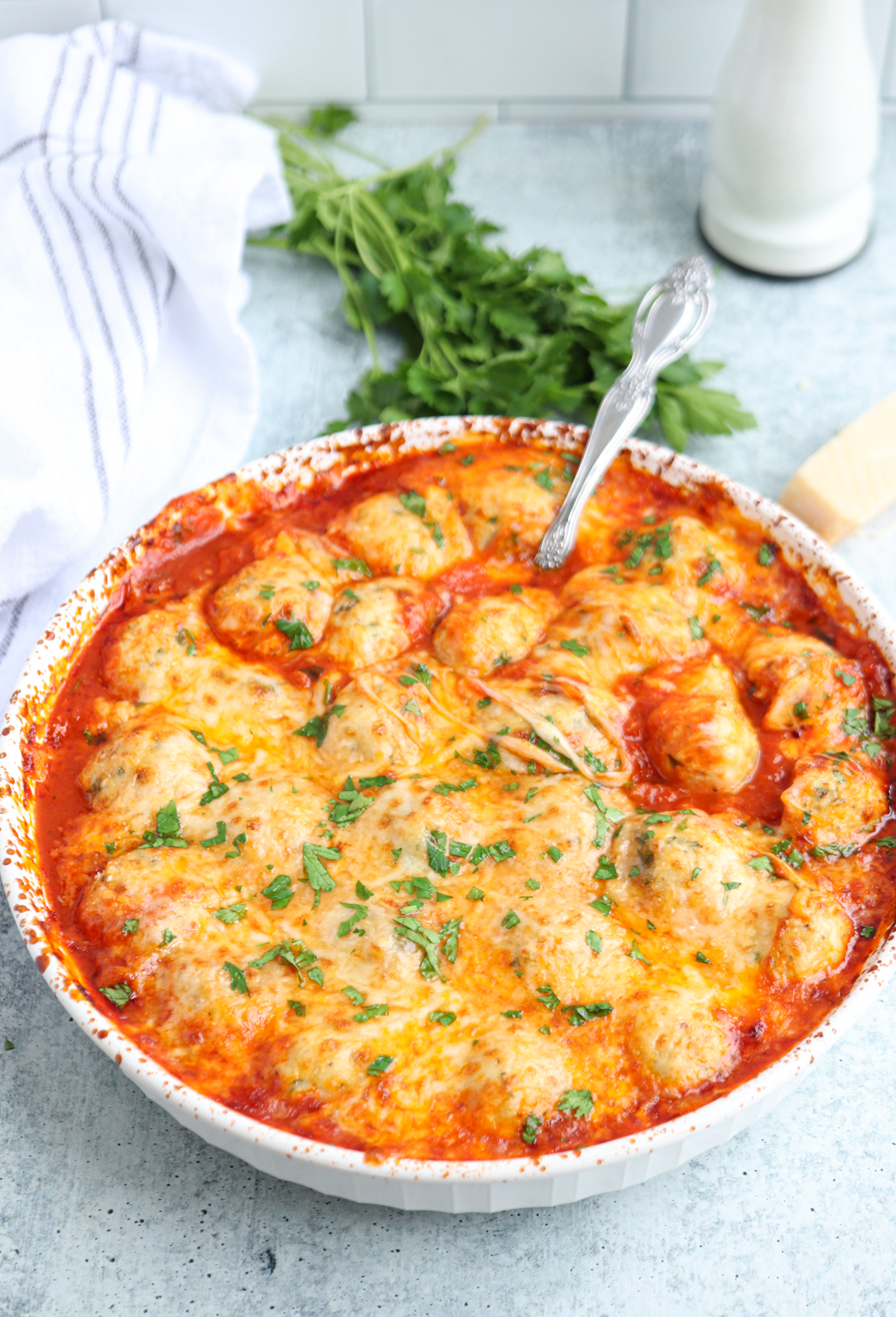 Baked dish of cheesy chicken parm meatballs in marinara sauce, topped with melted golden cheese and fresh chopped parsley, served in a white round baking dish on a gray surface with herbs, a towel, and a spoon in the background.