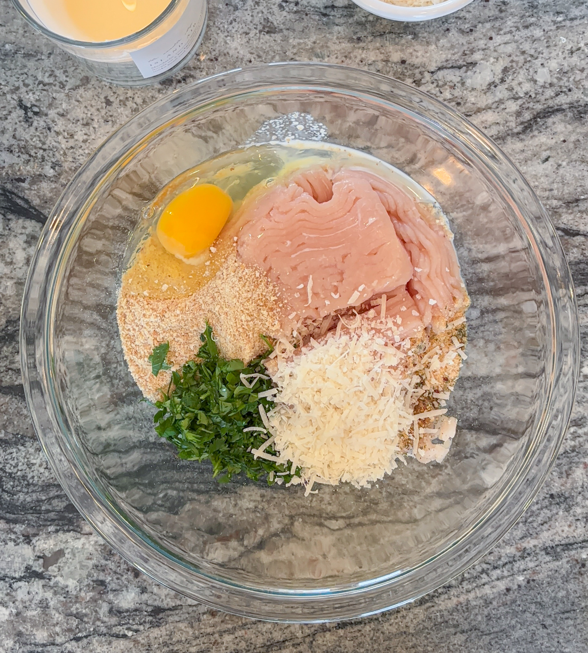 A glass bowl on a granite countertop contains ground chicken, an egg, breadcrumbs, chopped parsley, grated Parmesan cheese, and seasonings—ingredients ready to be mixed for delicious chicken parm meatballs.