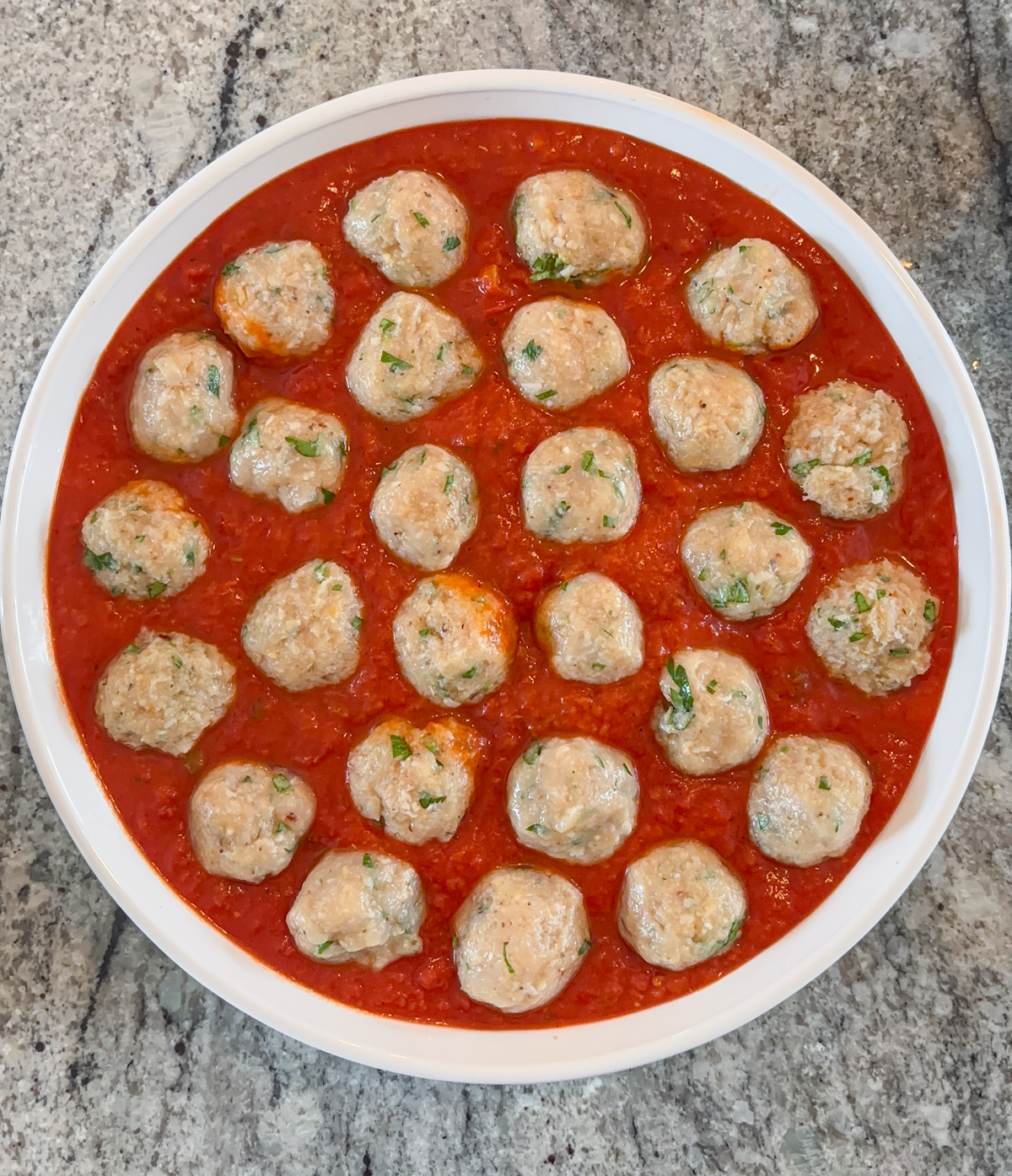 A white dish filled with chicken parm meatballs garnished with herbs, arranged in a circle and nestled in a bright red tomato sauce, set on a gray granite countertop.