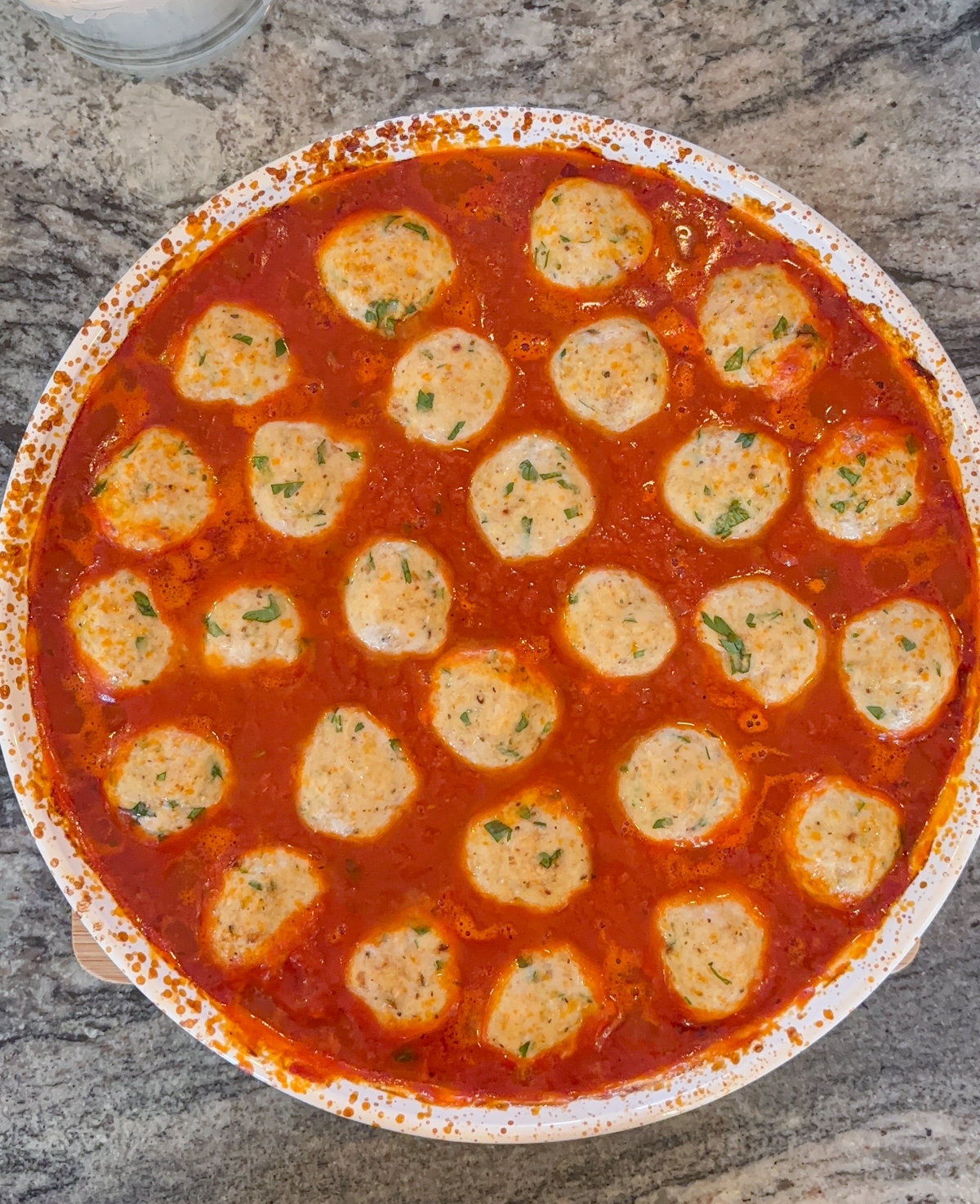 A white pan filled with chicken parm meatballs in a rich red tomato sauce, garnished with chopped herbs, sits on a marbled countertop.