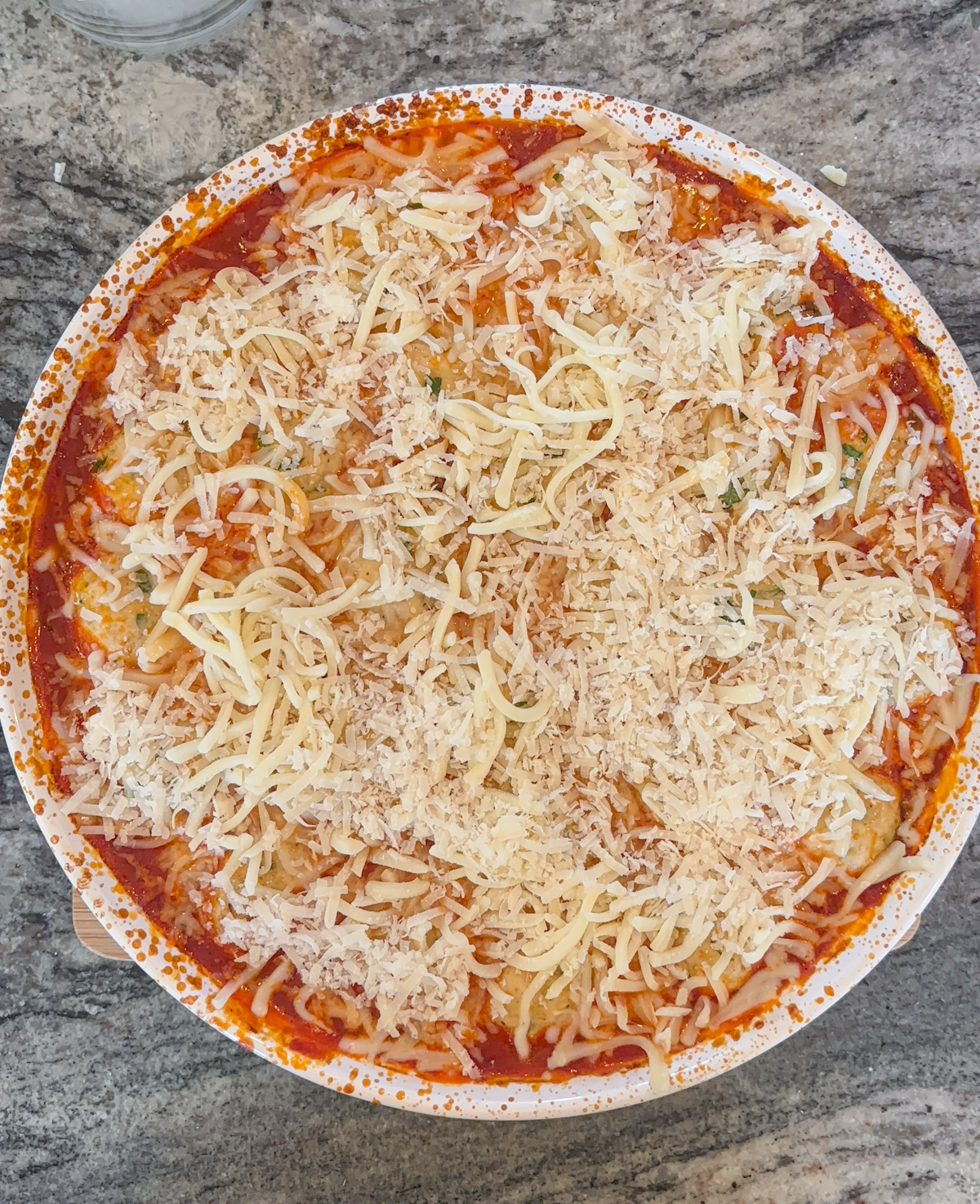 A round baking dish filled with pasta and chicken parm meatballs, covered in tomato sauce and generously topped with shredded cheese, placed on a gray marble countertop.