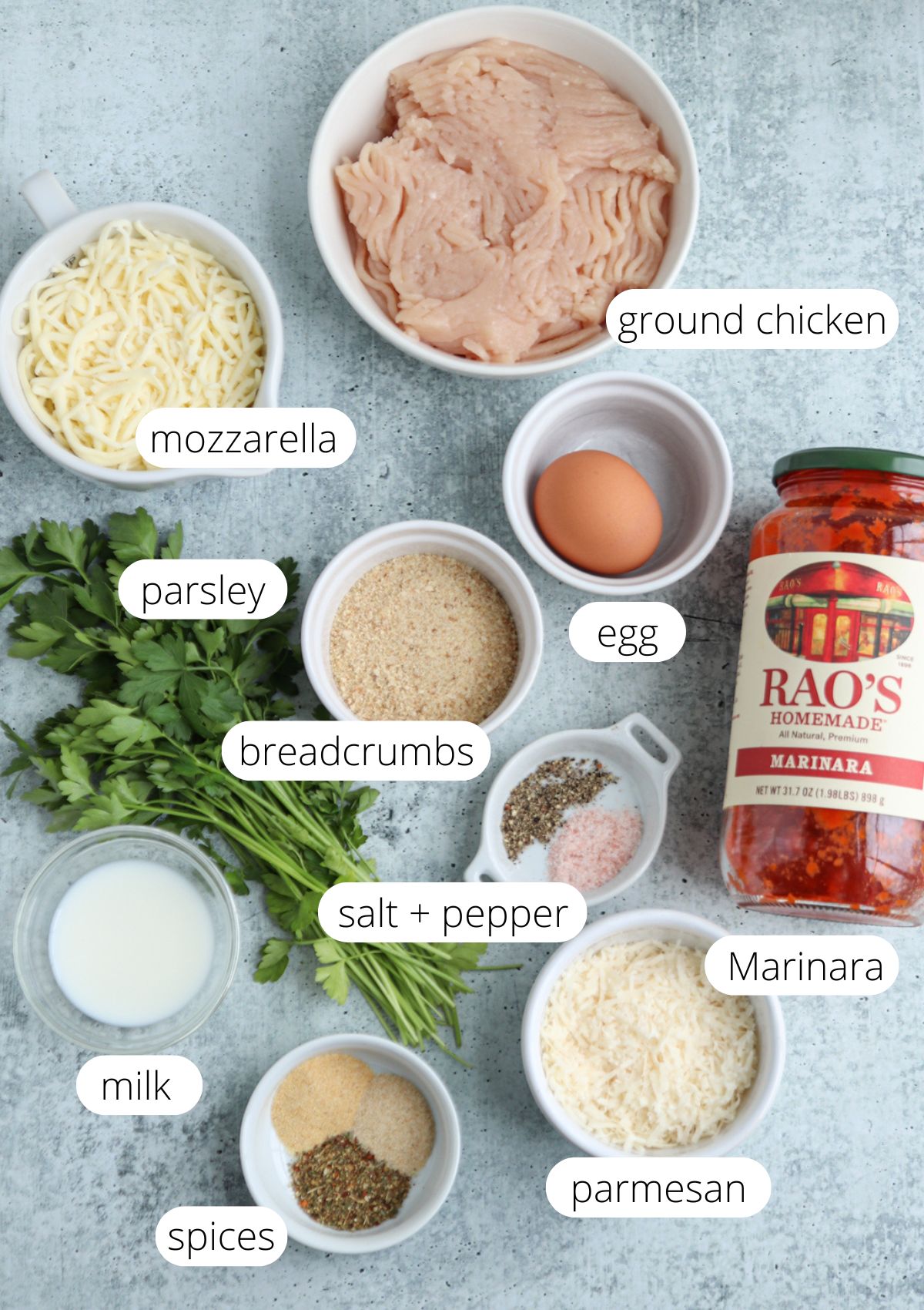 Ingredients for chicken parm meatballs, featuring ground chicken, mozzarella, parsley, breadcrumbs, egg, salt and pepper, marinara sauce, milk, spices and parmesan cheese—all labeled and arranged on a gray surface.