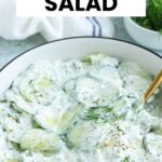 A white bowl filled with creamy Polish cucumber salad, garnished with fresh dill and a gold fork inside. Text over the image reads “Creamy Polish Cucumber Salad.”.