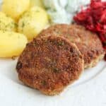Two browned kotlety mielone (Polish meat patties) are served on a white plate with boiled potatoes topped with dill, shredded beets, and a creamy salad in the background.