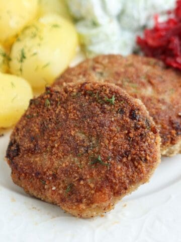 Two browned kotlety mielone (Polish meat patties) are served on a white plate with boiled potatoes topped with dill, shredded beets, and a creamy salad in the background.