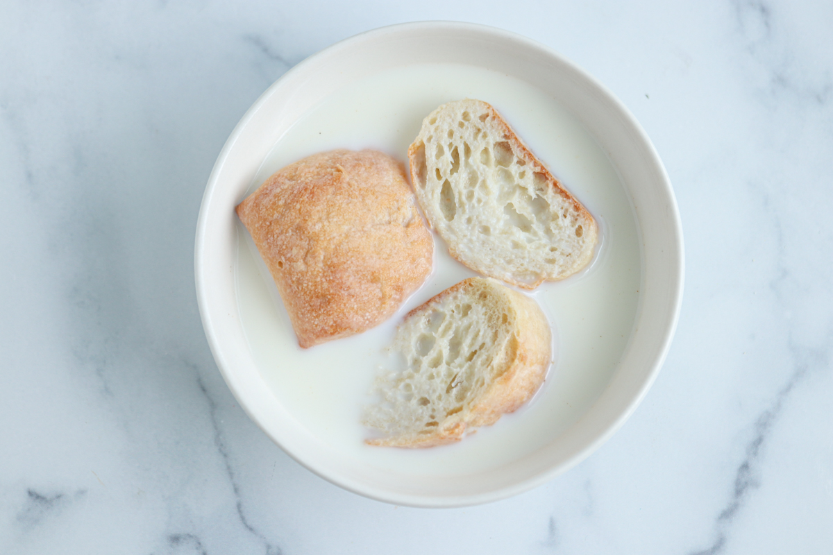 A white bowl filled with milk and three pieces of rustic bread, perfect for preparing kotlety mielone, placed on a light marble surface.