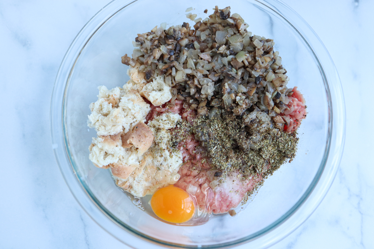 A glass bowl contains the classic kotlety mielone mixture—ground meat, chopped cooked onions and mushrooms, torn bread pieces, a raw egg, and dried herbs—all on a white surface.