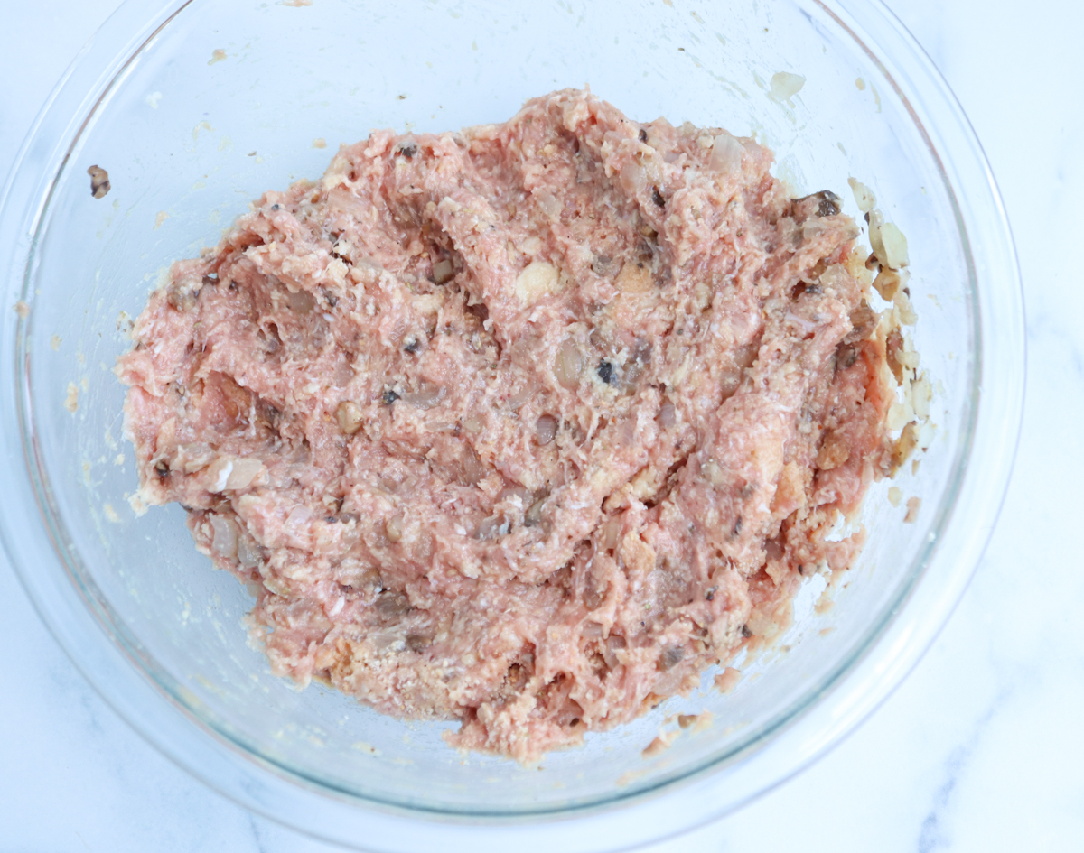A glass bowl filled with a mixture of ground meat, chopped onions, and seasonings sits on a white surface, ready to be shaped into classic kotlety mielone.