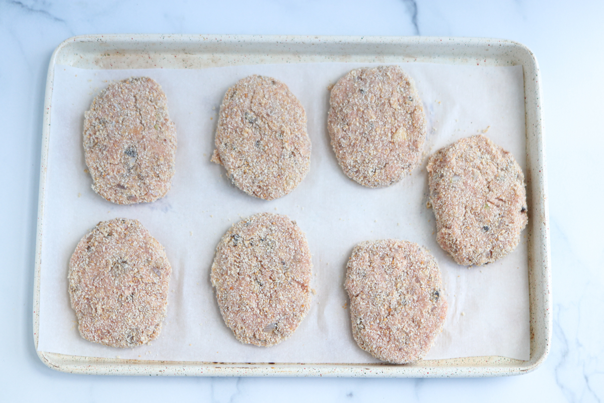 A baking sheet lined with parchment paper holds seven uncooked, breaded kotlety mielone spaced evenly apart on a light marble surface.