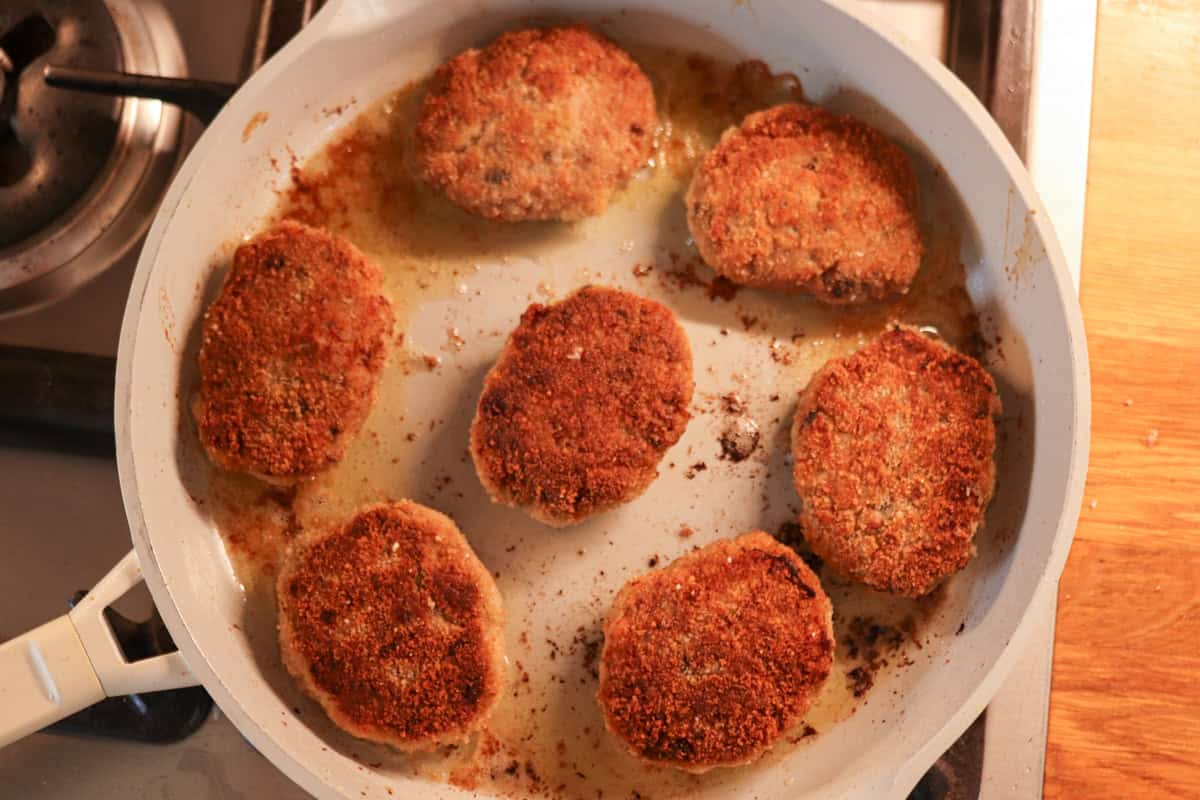 Seven breaded kotlety mielone are frying in a white skillet on a stove, showing a golden-brown, crispy exterior with some oil and browned bits in the pan.