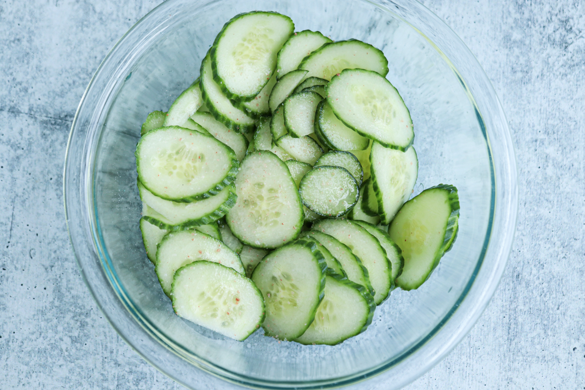 A glass bowl filled with thinly sliced cucumbers sprinkled with salt sits on a light gray textured surface, ready to become a refreshing Polish cucumber salad.