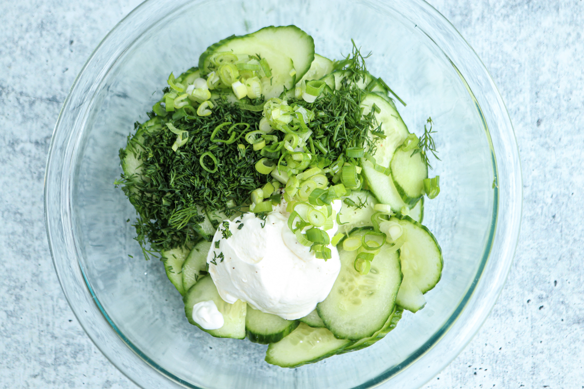 A glass bowl with sliced cucumbers, chopped green onions, fresh dill, and a dollop of sour cream or yogurt on a light textured surface, ready to be mixed into a classic Polish cucumber salad.