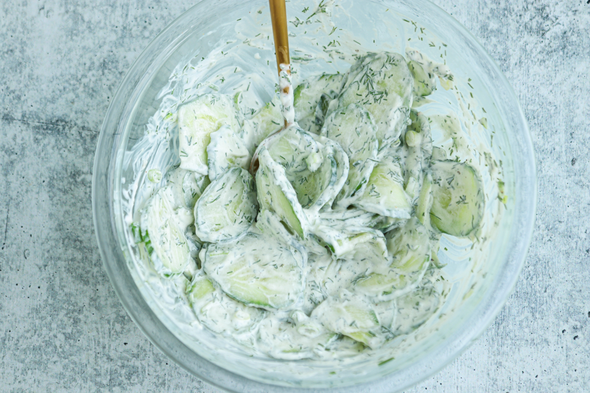 A glass bowl filled with creamy Polish cucumber salad, featuring cucumber slices coated in a thick, white dressing with herbs, and a spoon resting inside the bowl.