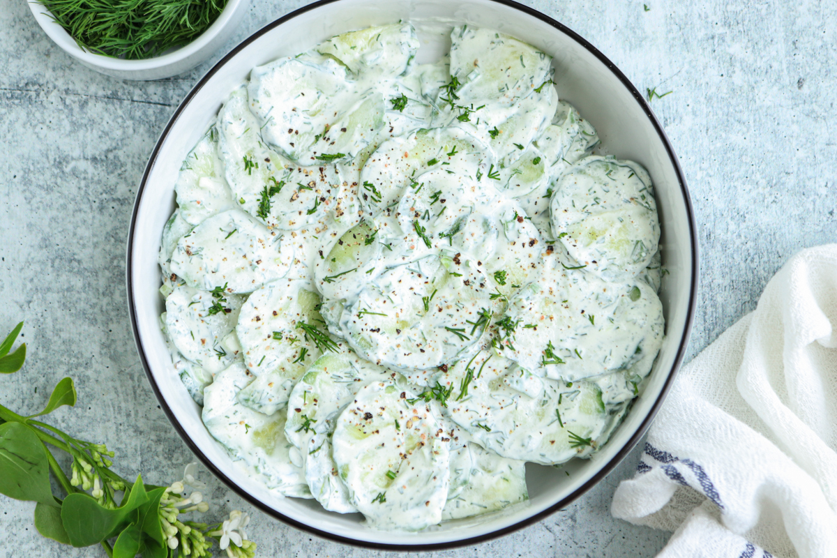 A bowl of creamy Polish cucumber salad garnished with fresh dill and ground pepper, placed on a light textured surface. A small bowl of dill and a white napkin are nearby.