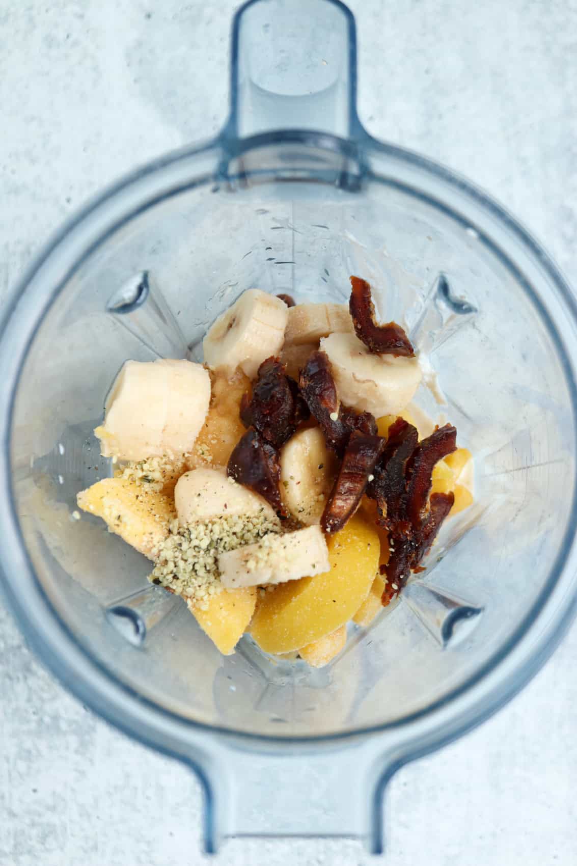 A top-down view of a blender containing sliced bananas, dates, pineapple chunks, and a sprinkle of seeds—perfect ingredients for a peach yogurt smoothie—ready to be blended on a light textured surface.