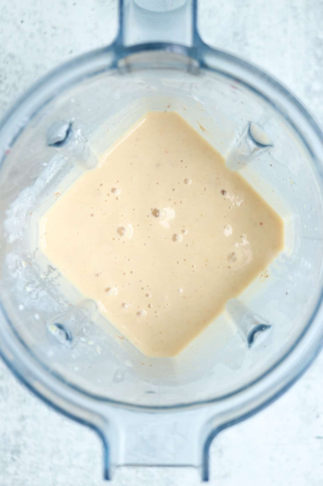 A creamy, light beige peach yogurt smoothie mixture with small bubbles is seen inside a blender viewed from above, against a light background.