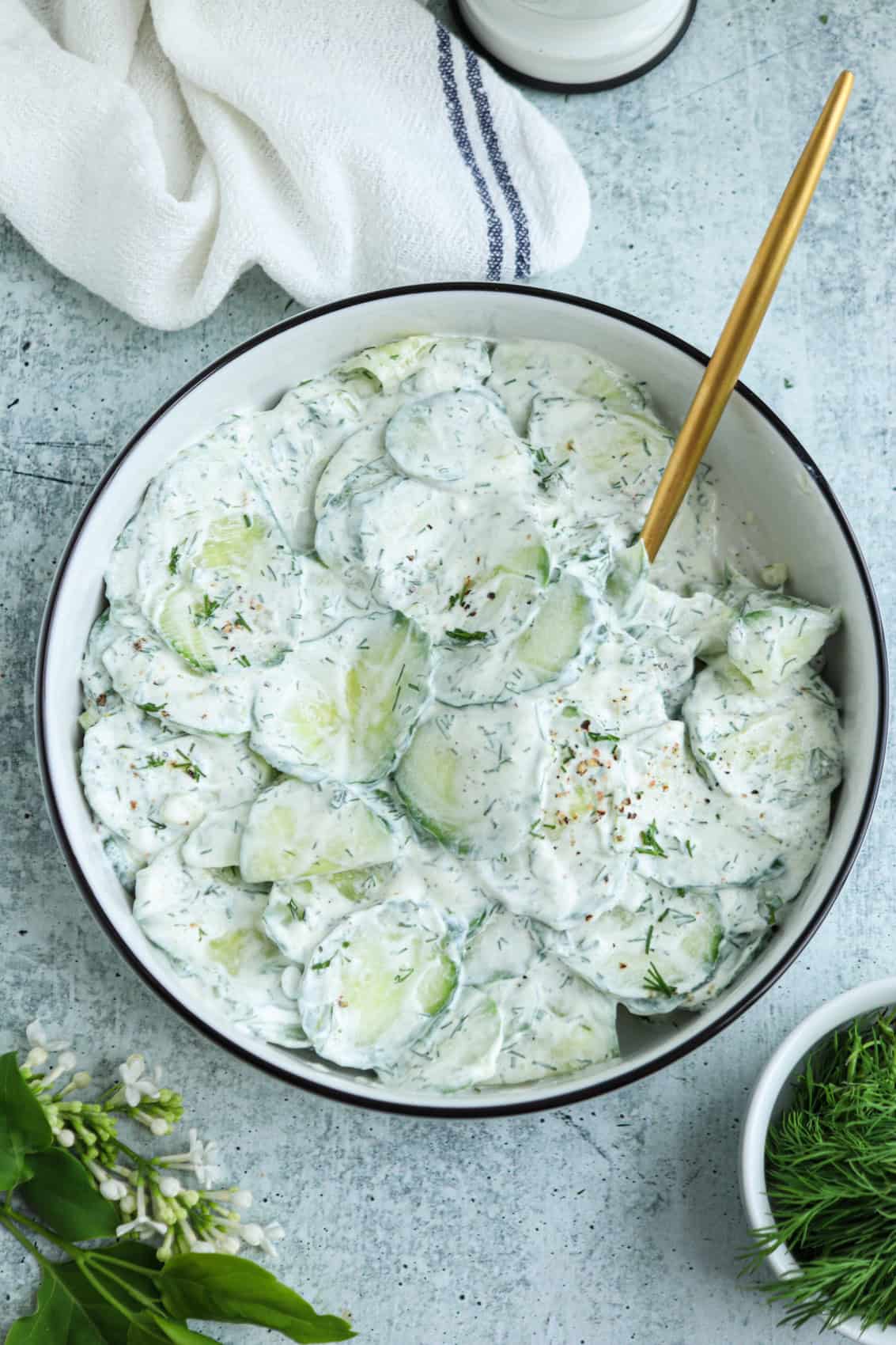A bowl of creamy Polish cucumber salad with fresh dill, sliced cucumbers, and a gold spoon, set on a light gray table with a white cloth and green herbs beside it.