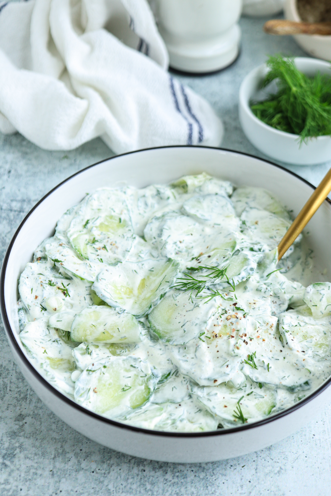 A bowl of creamy Polish cucumber salad with fresh dill, garnished with black pepper, sits on a light grey surface next to a small bowl of dill and a white cloth. A gold spoon rests in the salad.