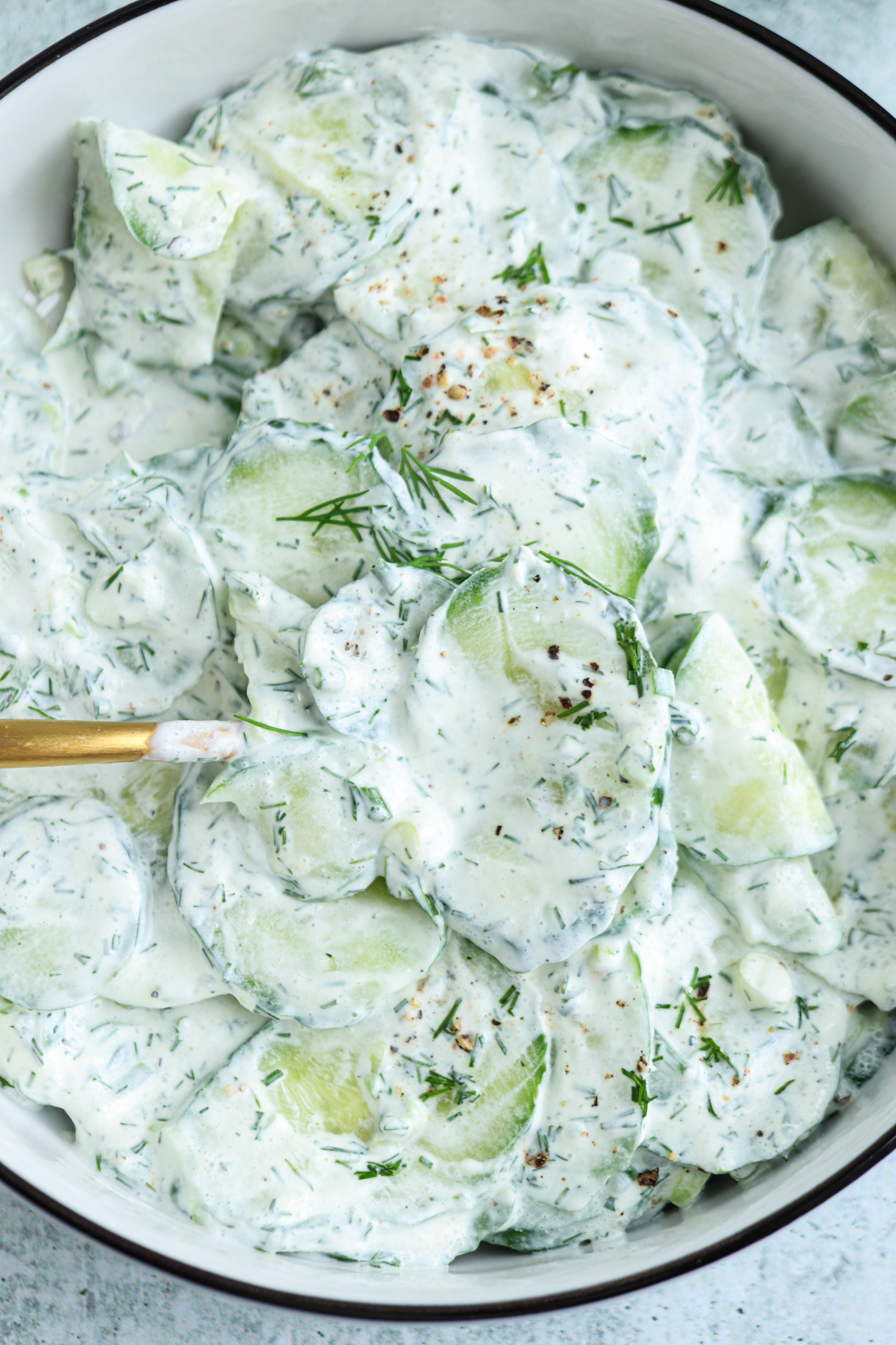 Bowl of Polish cucumber salad with creamy dressing, garnished with fresh dill and cracked pepper. A gold fork is partially visible on the left side.