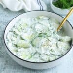 A white bowl filled with creamy Polish cucumber salad garnished with fresh dill, sitting on a light textured surface with a gold spoon and a small bowl of dill in the background.