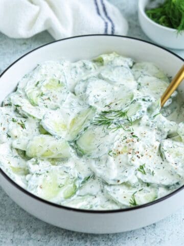 A white bowl filled with creamy Polish cucumber salad garnished with fresh dill, sitting on a light textured surface with a gold spoon and a small bowl of dill in the background.