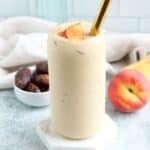 A creamy peach yogurt smoothie in a tall glass, topped with peach slices and a sprinkle of cinnamon. A gold straw rests in the glass. Nearby are a fresh peach and a bowl of dates, with a light cloth in the background.